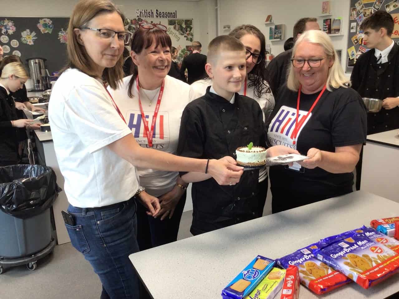The runner up student stands with Hill Biscuits staff and holds his cheesecake at Laurus Ryecroft on National Biscuit Day.